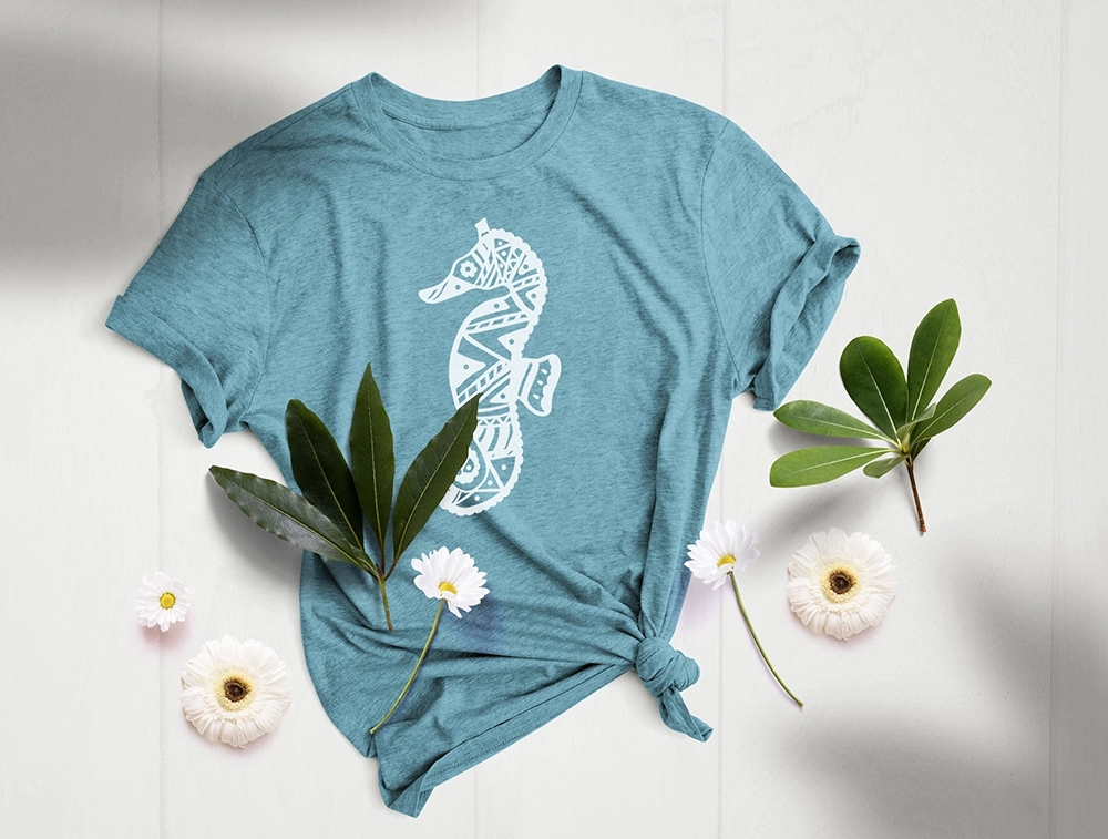 Seepferdchen Bügelbild – Weißes Mandala-Motiv auf blauem T-Shirt im floralen Flatlay Blaues T-Shirt mit weißem Seepferdchen-Bügelbild im Mandala-Stil, dekorativ arrangiert mit Blättern und weißen Blumen auf hellem Untergrund